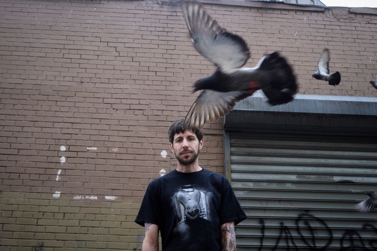 Underground hip-hop artist GDP wearing a graphic t-shirt in front of a brick wall as pigeons fly past, photographed by Rochelle Marie Adam.